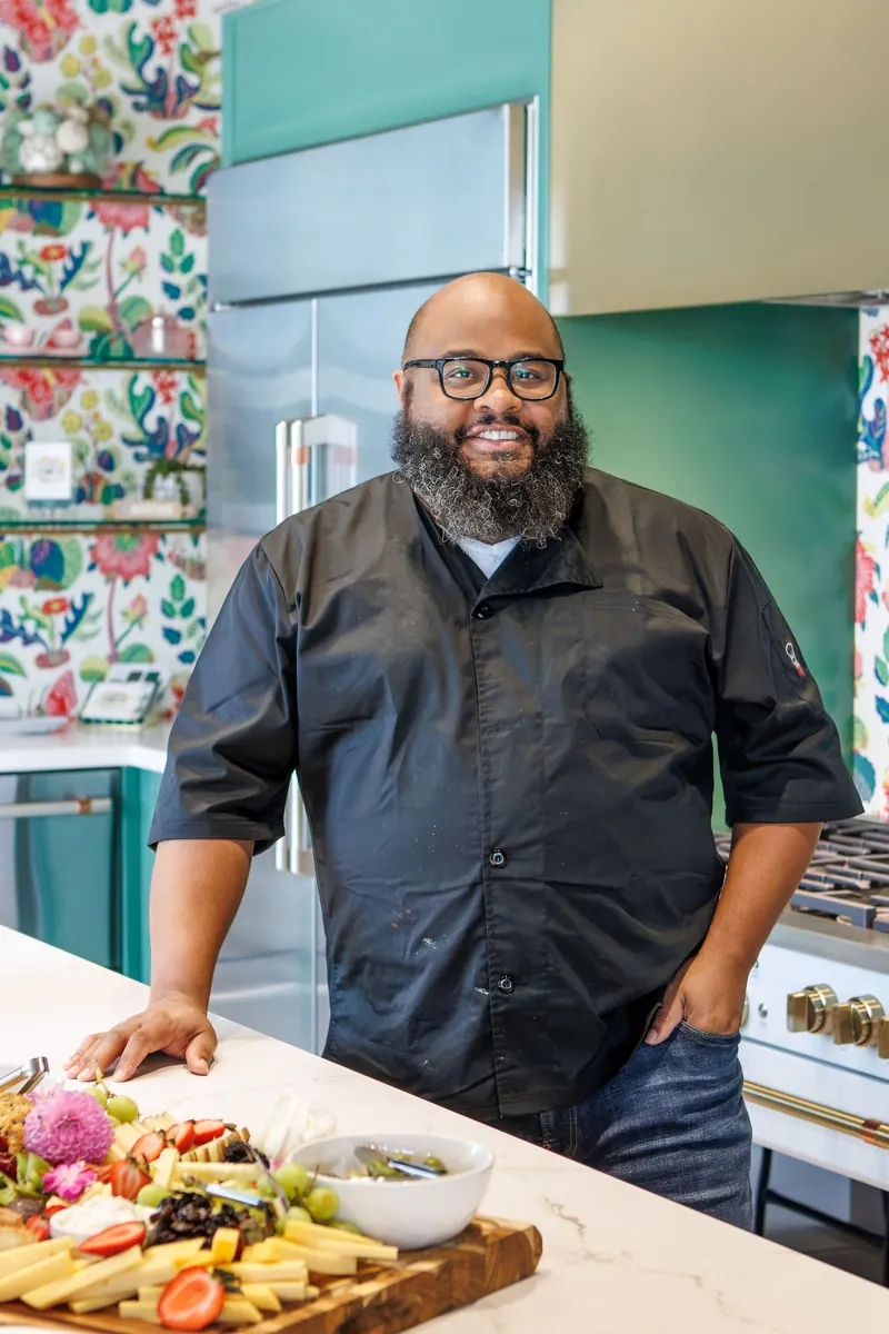 A smiling chef in a black chef coat leans on a marble island set with a charcuterie board, green cabinets and floral wallpaper behind him