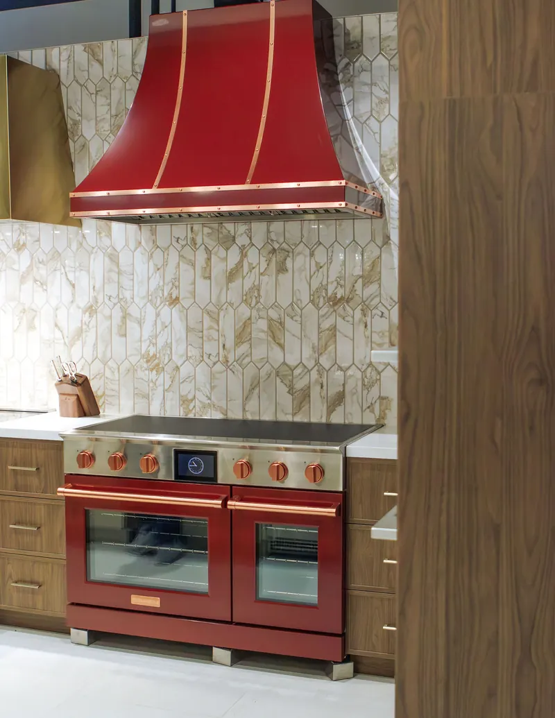A glossy red range and copper hood set against patterned green tile in an appliance showroom