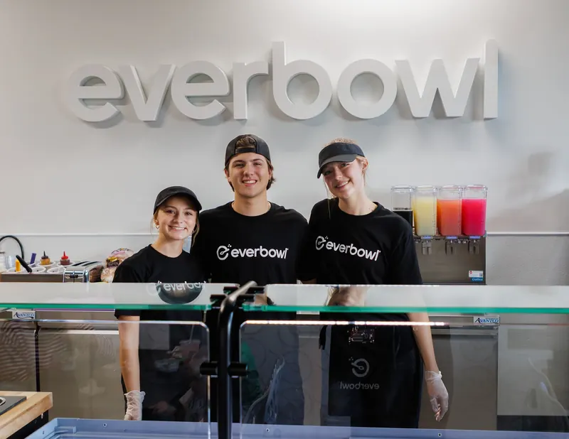 Three Everbowl crew members in black tees and visors smile behind the counter under the Everbowl sign