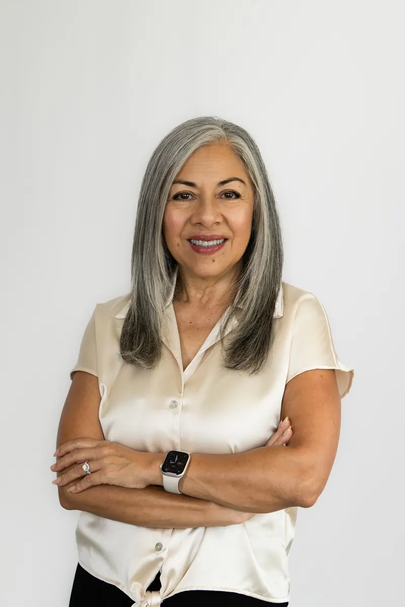 A founder with long silver hair and a cream tie-front blouse smiles with her arms crossed against a white backdrop