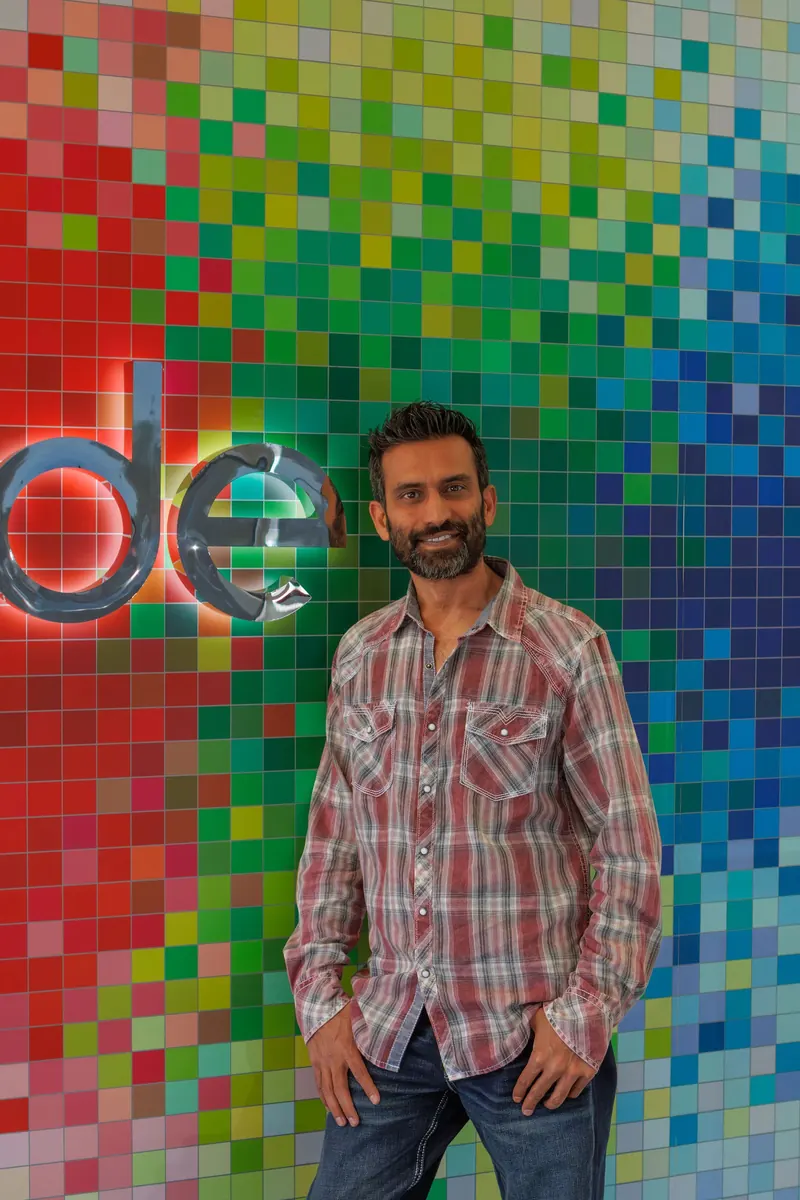 A coding school owner in a plaid shirt stands in front of a bright rainbow pixel wall with the iCode logo