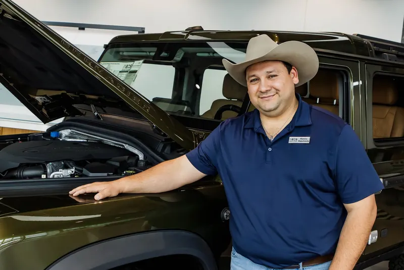 A dealership salesman in a cowboy hat and navy polo leans on the open hood of an olive Grenadier