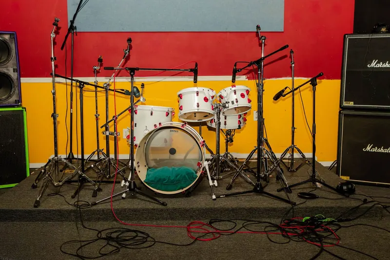 A drum kit set up in a recording studio live room painted red and yellow, with amps along the wall