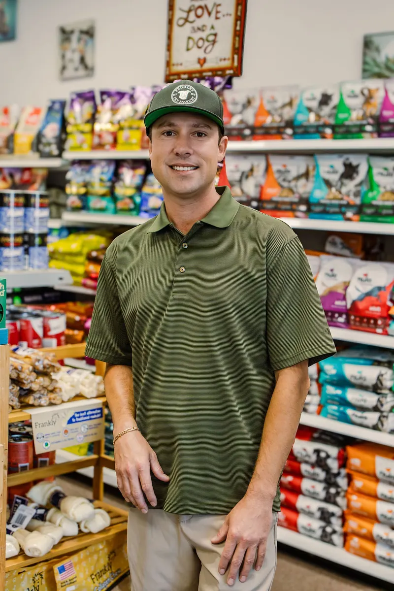 A pet store owner in a green polo and cap stands in an aisle of colorful pet food bags, smiling