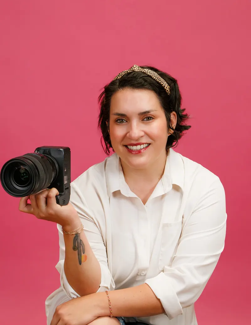A photographer in a white shirt sits on a wood floor against a hot pink backdrop, holding her camera and smiling