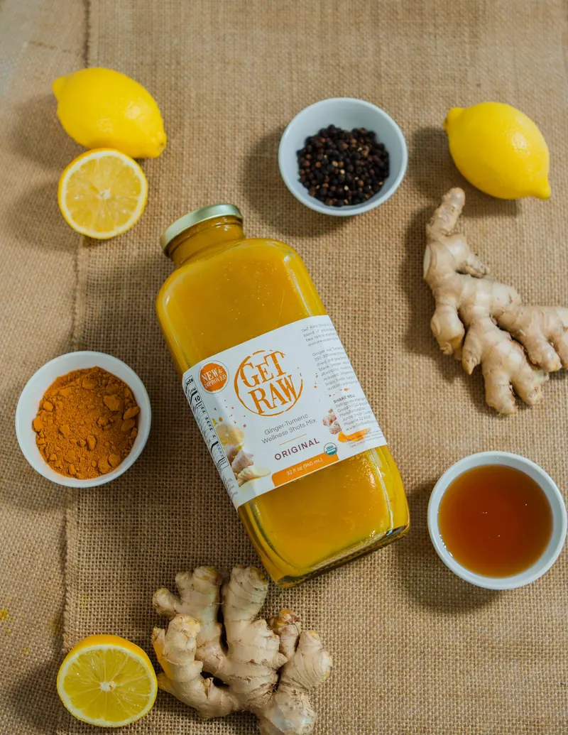 Overhead flat lay of a Get Raw ginger turmeric bottle on burlap with lemons, fresh ginger and small bowls of turmeric and pepper