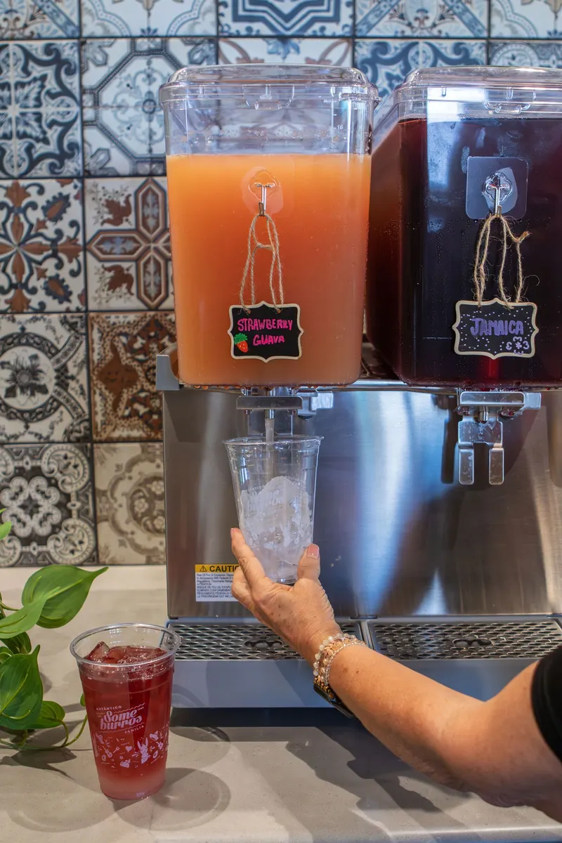 A hand fills a cup from an agua fresca dispenser against a tiled wall