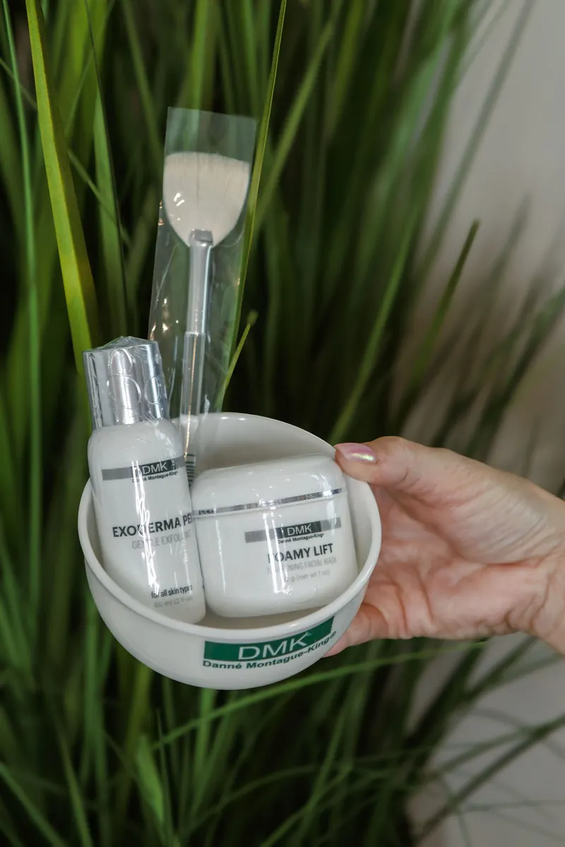 A hand holds a skincare jar beside a green fern