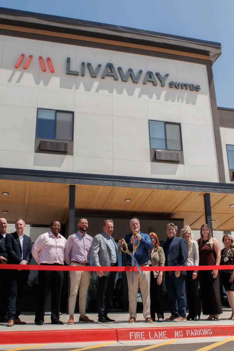 The LivAway Suites team and guests line up behind a red ribbon with giant scissors under the new building's sign