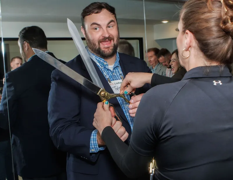 A guest poses with giant ribbon scissors while others laugh nearby