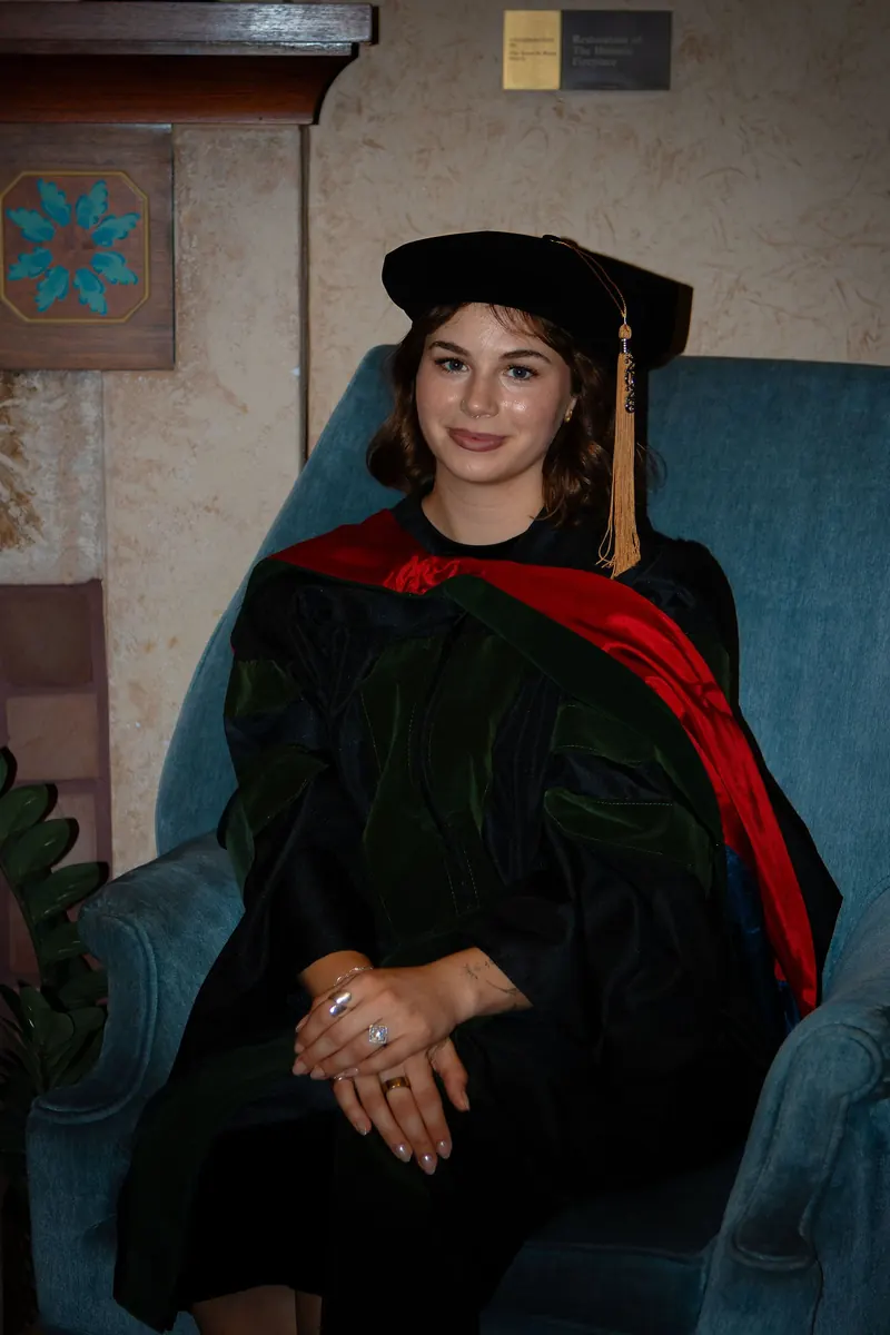 A new doctor in a black doctoral gown with a red hood sits in a blue velvet armchair