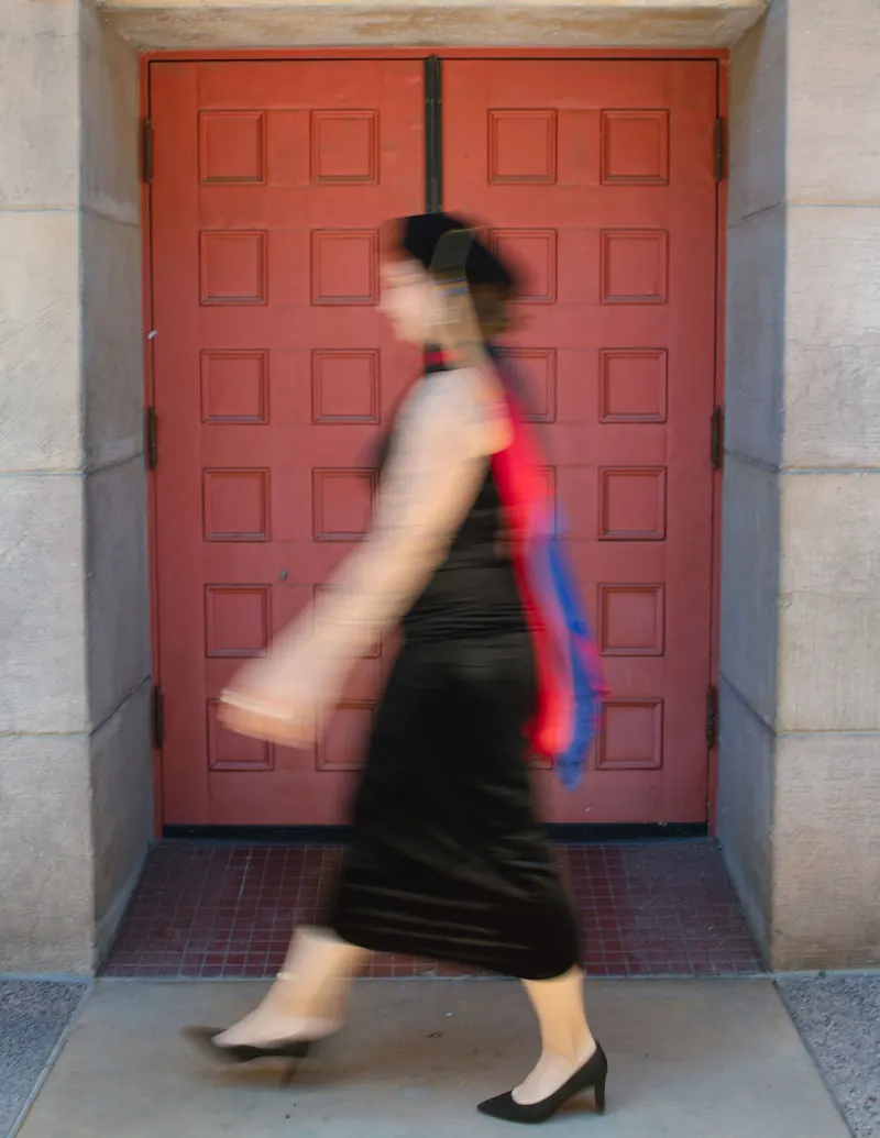 A graduate in a black dress and red sash walks past tall red doors, her stride softly blurred