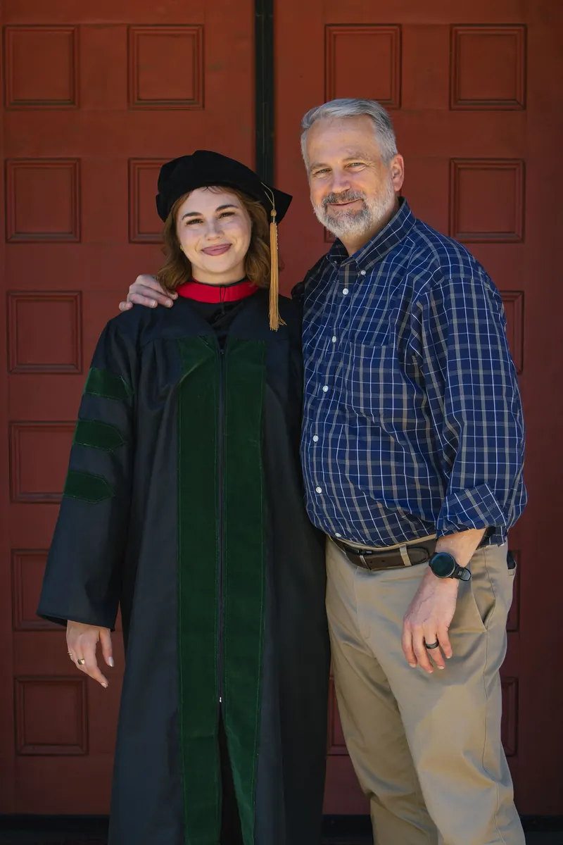 A doctoral graduate and her father smile together in front of tall red doors