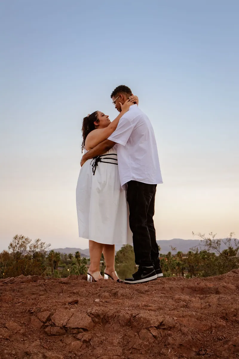 An engaged couple stands on red desert rock under a wide blue sky