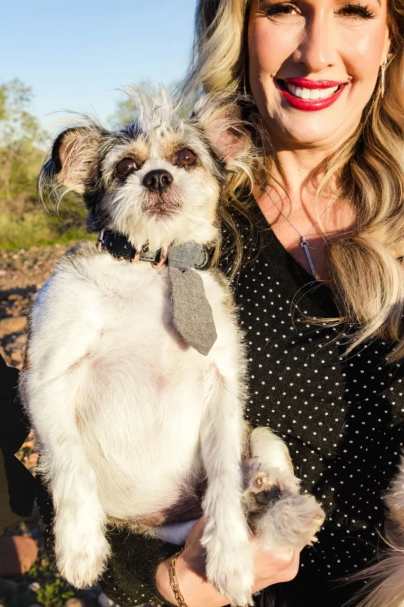 A woman in a black dress laughs while holding her shih tzu in the desert