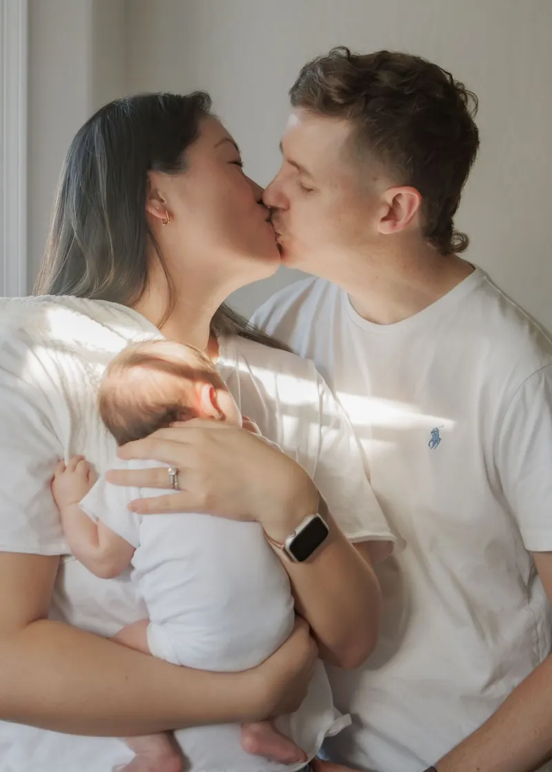 New parents share a kiss over their sleeping newborn in soft window light