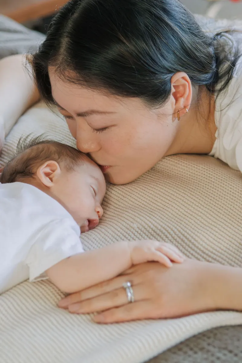 A mom lies beside her sleeping newborn and kisses her cheek