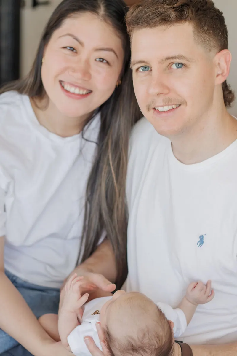 New parents in white tees smile on their bed with their newborn