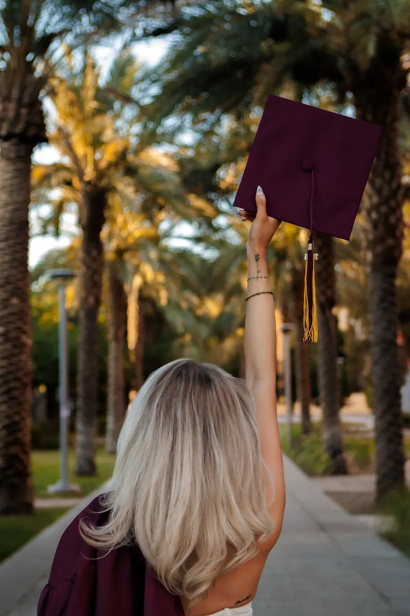 A graduate seen from behind tosses her cap into the air on a palm-lined walk