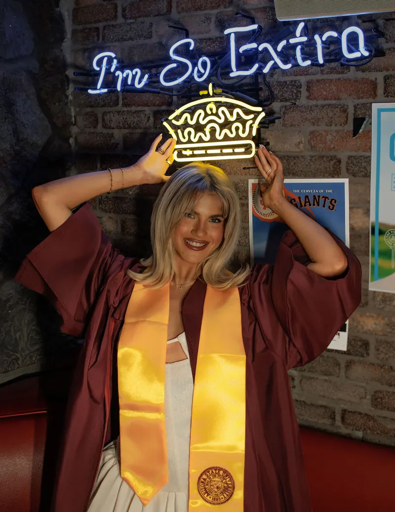 A graduate in a maroon gown wears a sparkly crown under a pink neon sign