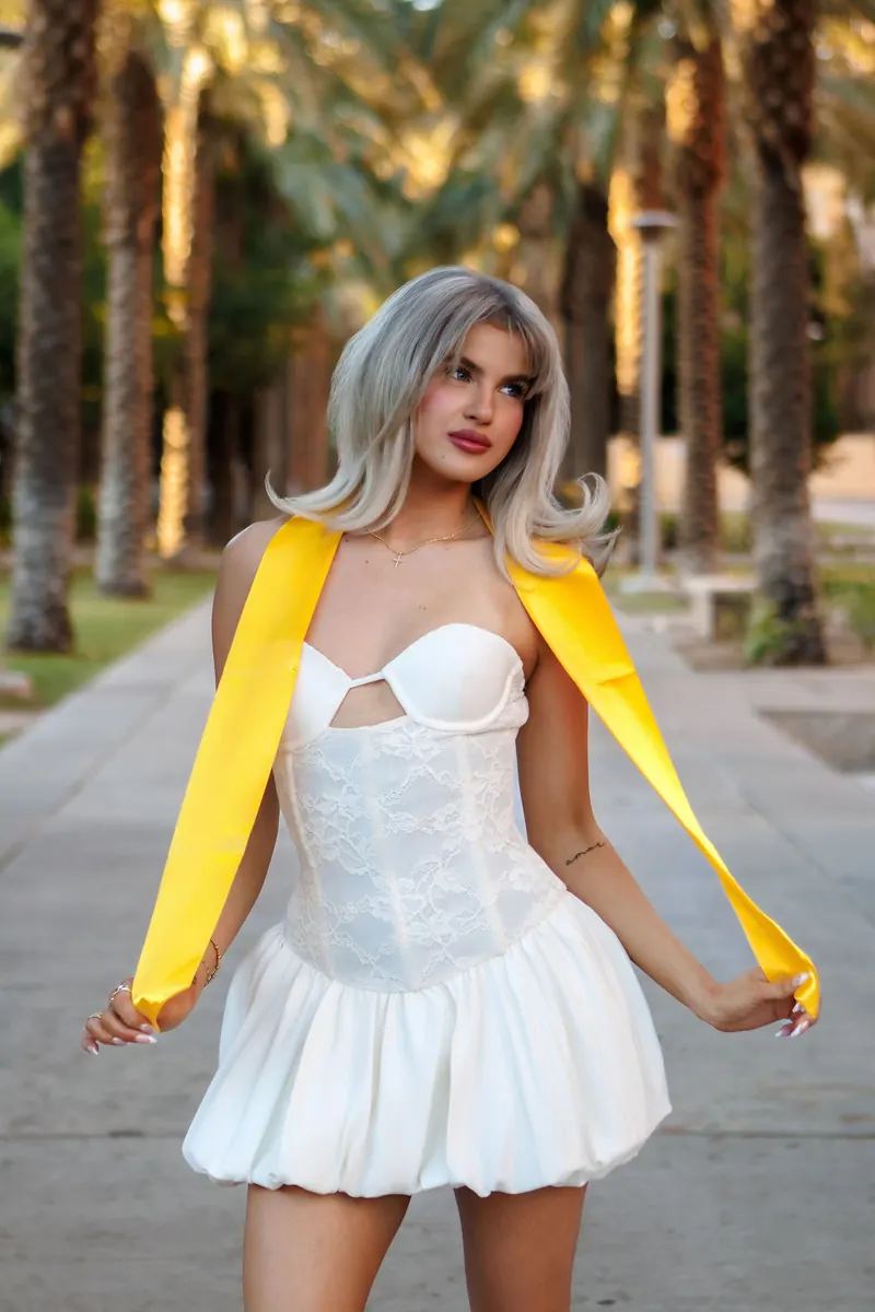 A graduate in a white dress with a gold stole draped over her shoulders, standing on a palm-lined campus walk at golden hour