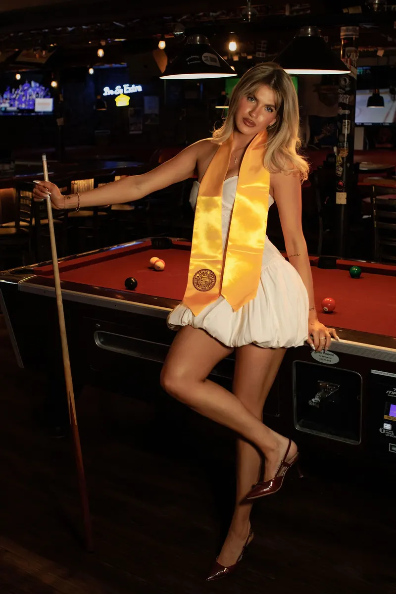 A graduate in a maroon gown and gold stole perches on a red pool table in a moody sports bar