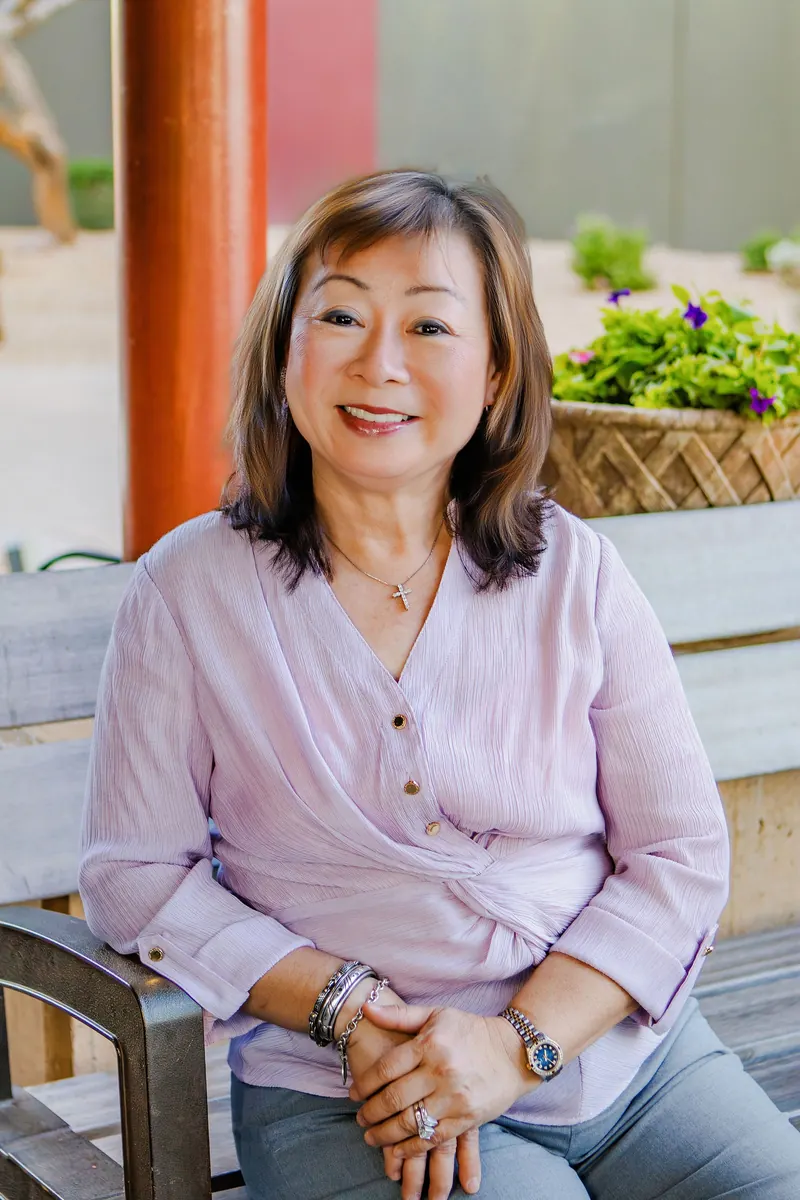 A smiling woman in a lavender blouse sits on a patio bench