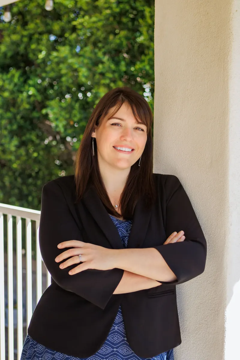A woman in a black blazer smiles with arms crossed on a white porch