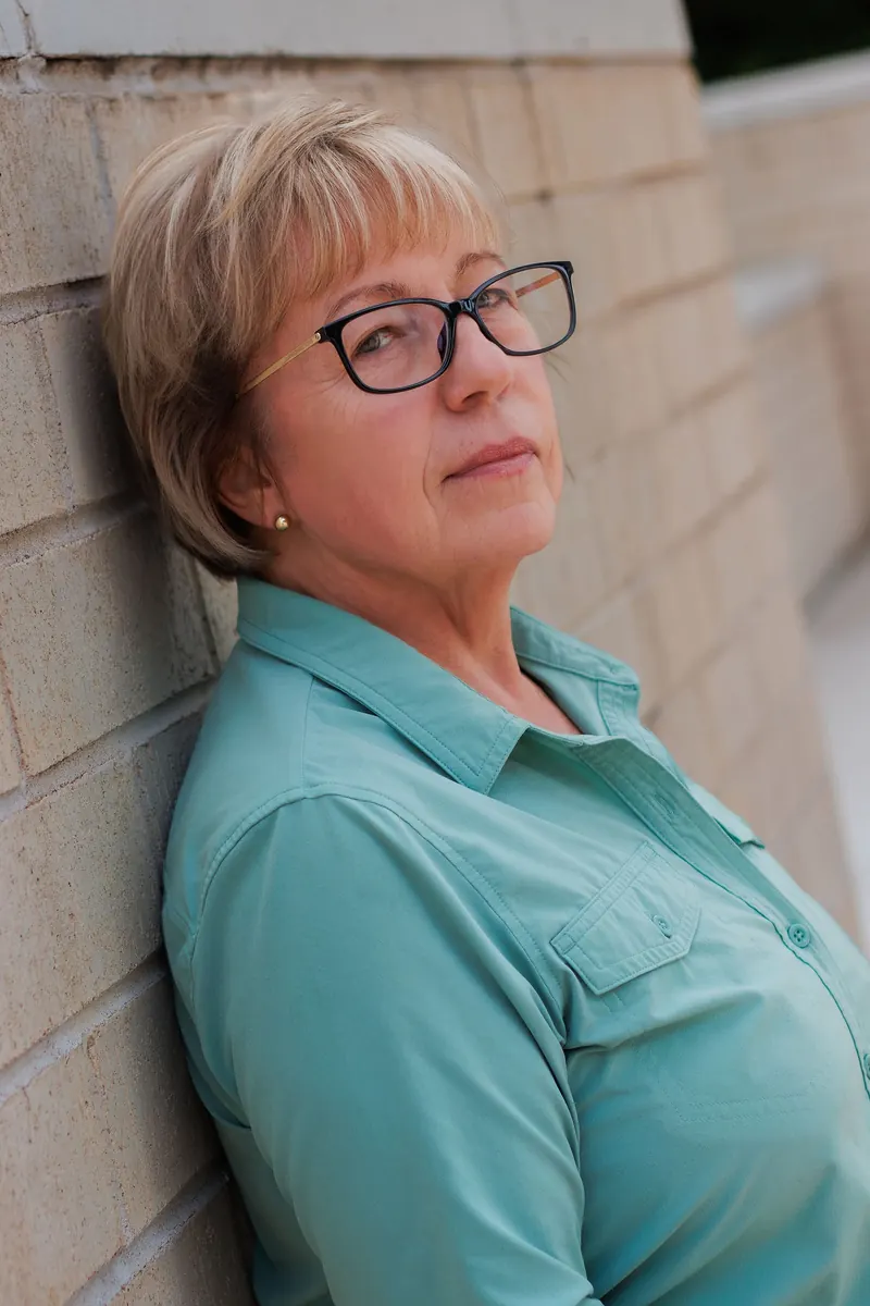 A woman in glasses and a teal shirt leans against a stucco wall